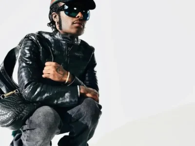 Lil Uzi Vert crouching in a black textured leather jacket, dark jeans, oversized sunglasses, and a black cap, carrying a matching black leather bag against a white background