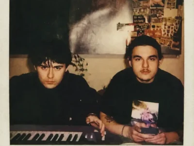 Vintage Polaroid-style photo of right Judah Weston and left IsoKeys men seated at a keyboard in a dim room, surround by posters and collage-covered walls, with warm muted tones, soft grain, and an aged off-white instant-film border