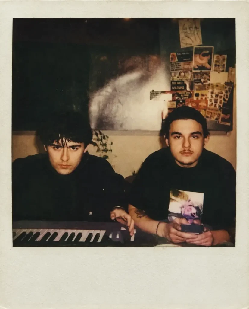 Vintage Polaroid-style photo of right Judah Weston and left IsoKeys men seated at a keyboard in a dim room, surround by posters and collage-covered walls, with warm muted tones, soft grain, and an aged off-white instant-film border