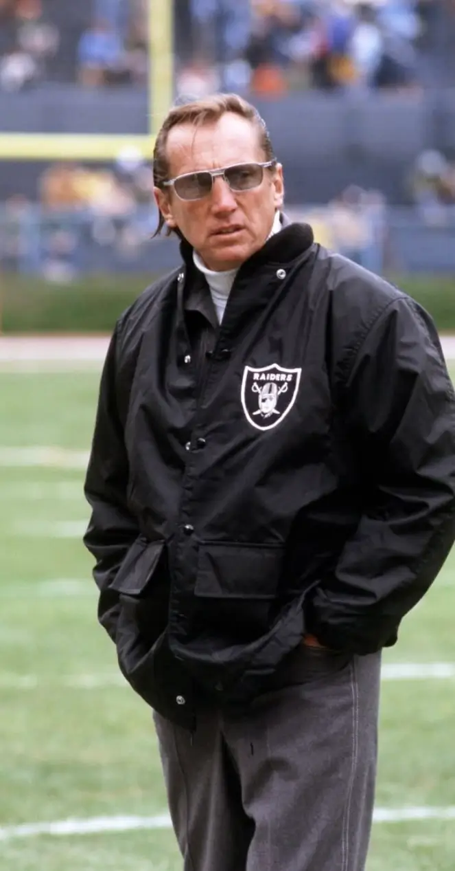 Man wearing tinted sunglasses and a black Raiders jacket standing on a football field inside a stadium.