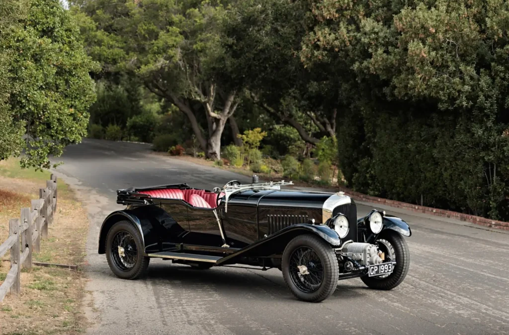 Vintage black open-top haute car with red leather seats, chrome details, wire-spoke wheels, and large round headlights parked on a winding tree-lined road