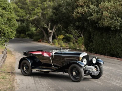 Vintage black open-top haute car with red leather seats, chrome details, wire-spoke wheels, and large round headlights parked on a winding tree-lined road