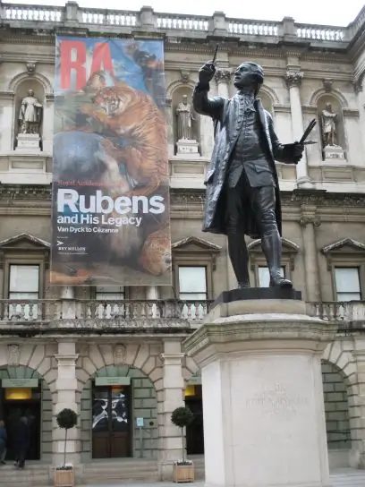 Bronze statue standing before a grand neoclassical museum facade featuring a large Rubens and His Legacy exhibition banner with a tiger artwork.
