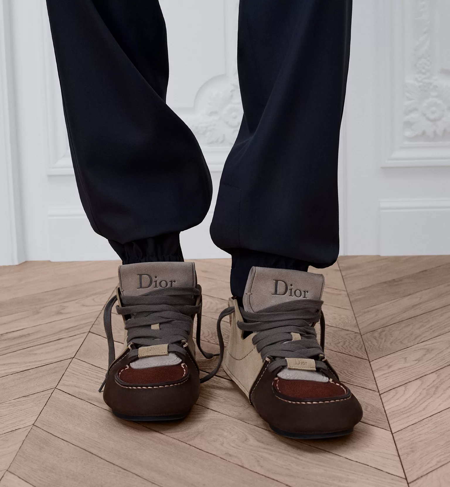 Dior shoes in brown and beige worn with black trousers on a parquet floor.