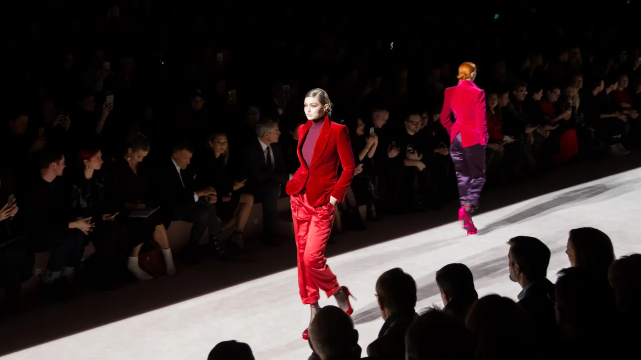 Models walk the runway in vibrant crimson tailoring during a fashion show, illuminated by dramatic spotlighting against a dark audience backdrop.