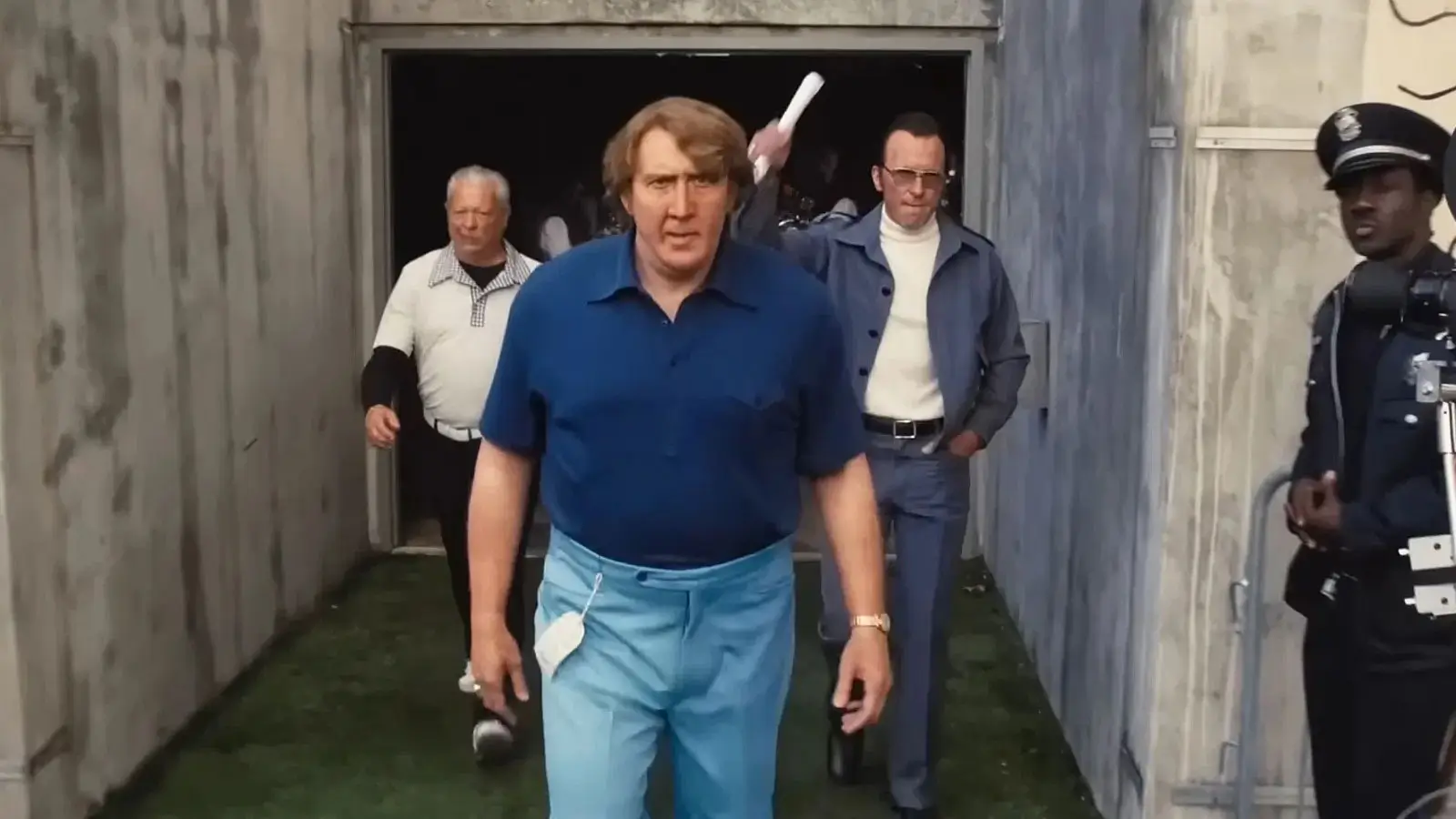 Man in a navy polo shirt and light blue pants walking out of a stadium tunnel, followed by several men and a uniformed police officer.