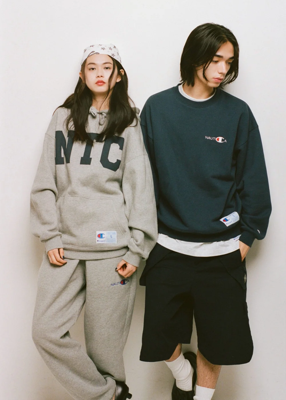 Two Models Wearing Champion X Nautica Apparel, Featuring A Gray NTC Graphic Hoodie And Sweatpants Alongside A Navy Nautica Crewneck Sweatshirt And Black Shorts.