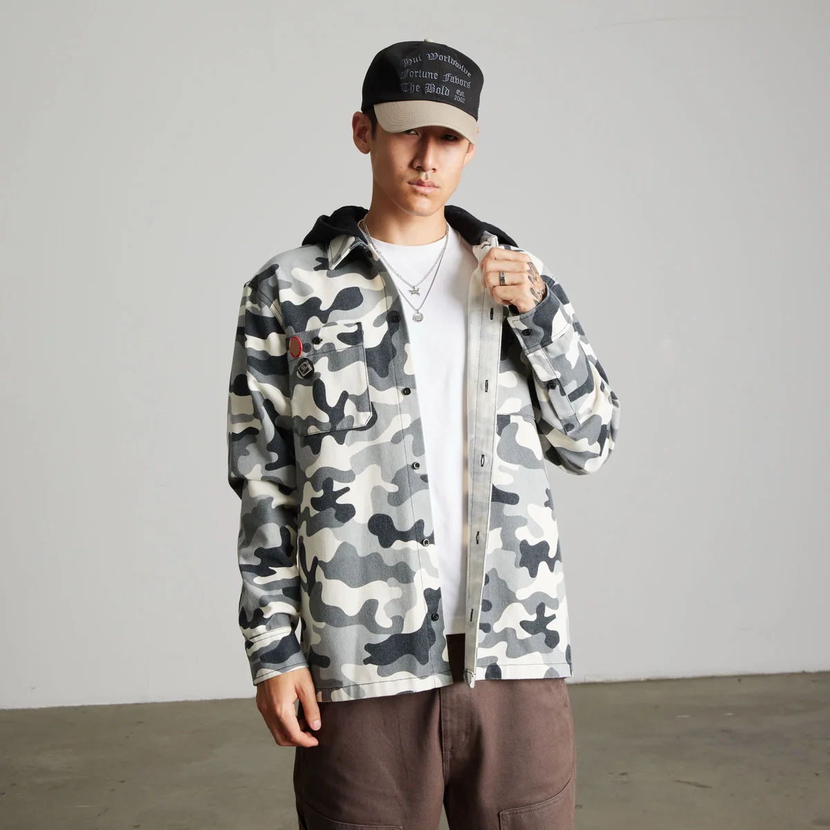 Model Wearing HUF Olympia Camo Hooded Overshirt Layered Over A White T-Shirt With Brown Trousers And A Two-Tone Graphic Cap