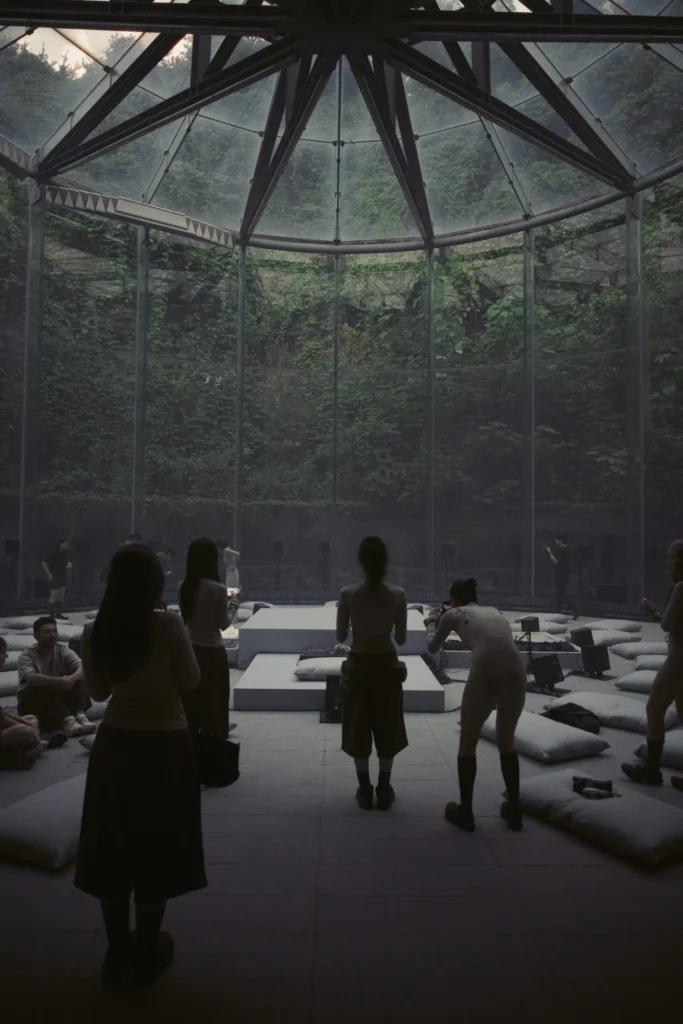 Visitors Gather Around A Min Show Install Inside A Circular Glass Pavilion Surround By Greenery