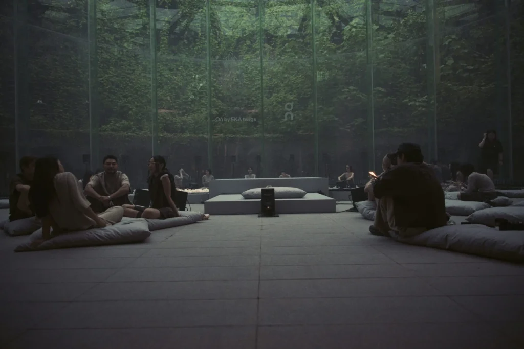 Guests Seat On Floor Cushion Inside A Glass-Wall Perform Space Surround By Dense Greenery