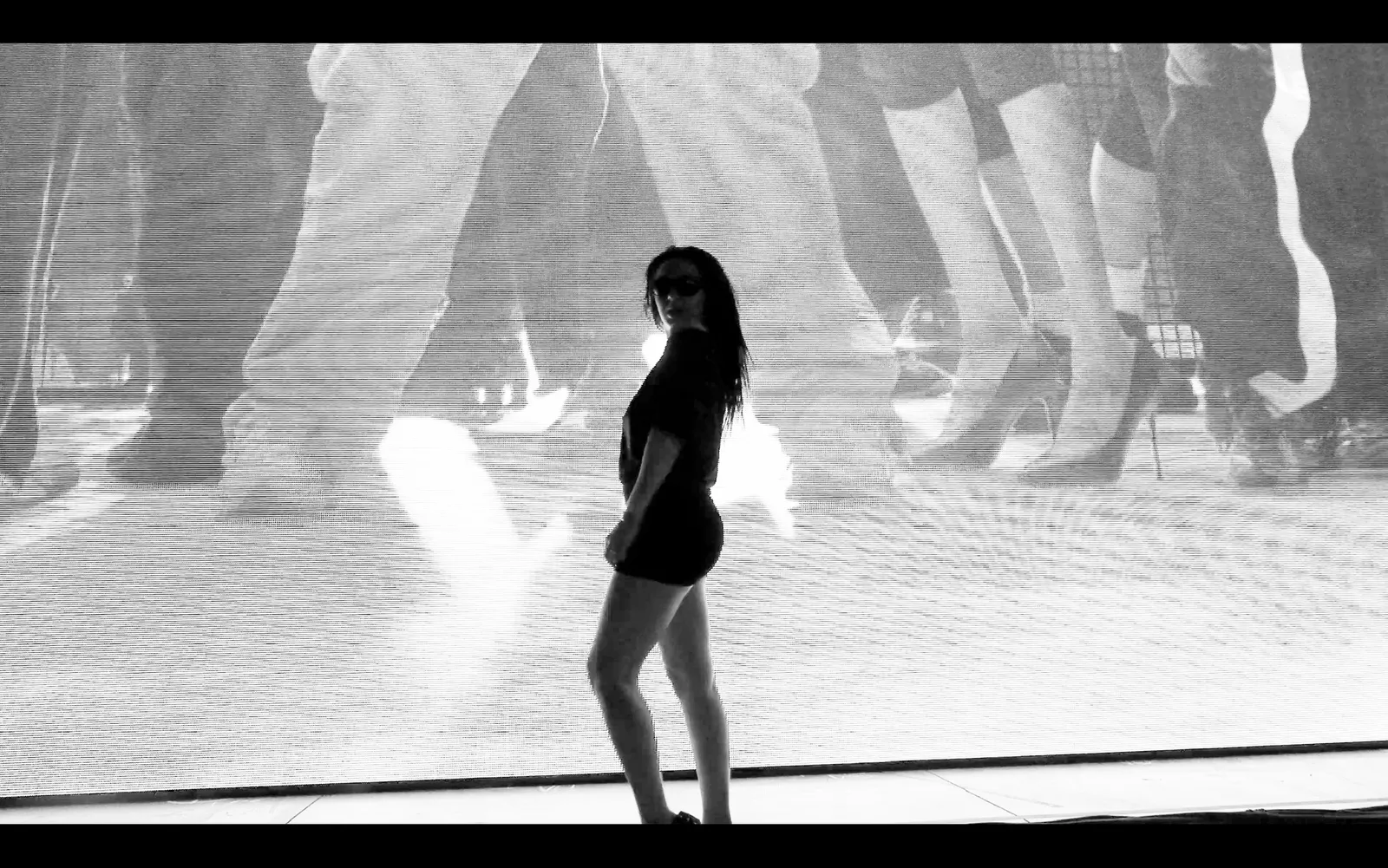 Black-and-white image of a woman in sunglasses posing onstage in front of a large video screen.