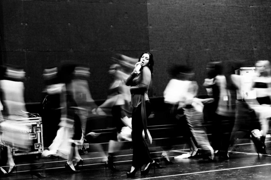 Black-and-white photograph of a female performer singing into a microphone at center stage as blurred dancers move rapidly around her during rehearsal