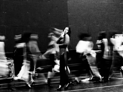 Black-and-white photograph of a female performer singing into a microphone at center stage as blurred dancers move rapidly around her during rehearsal