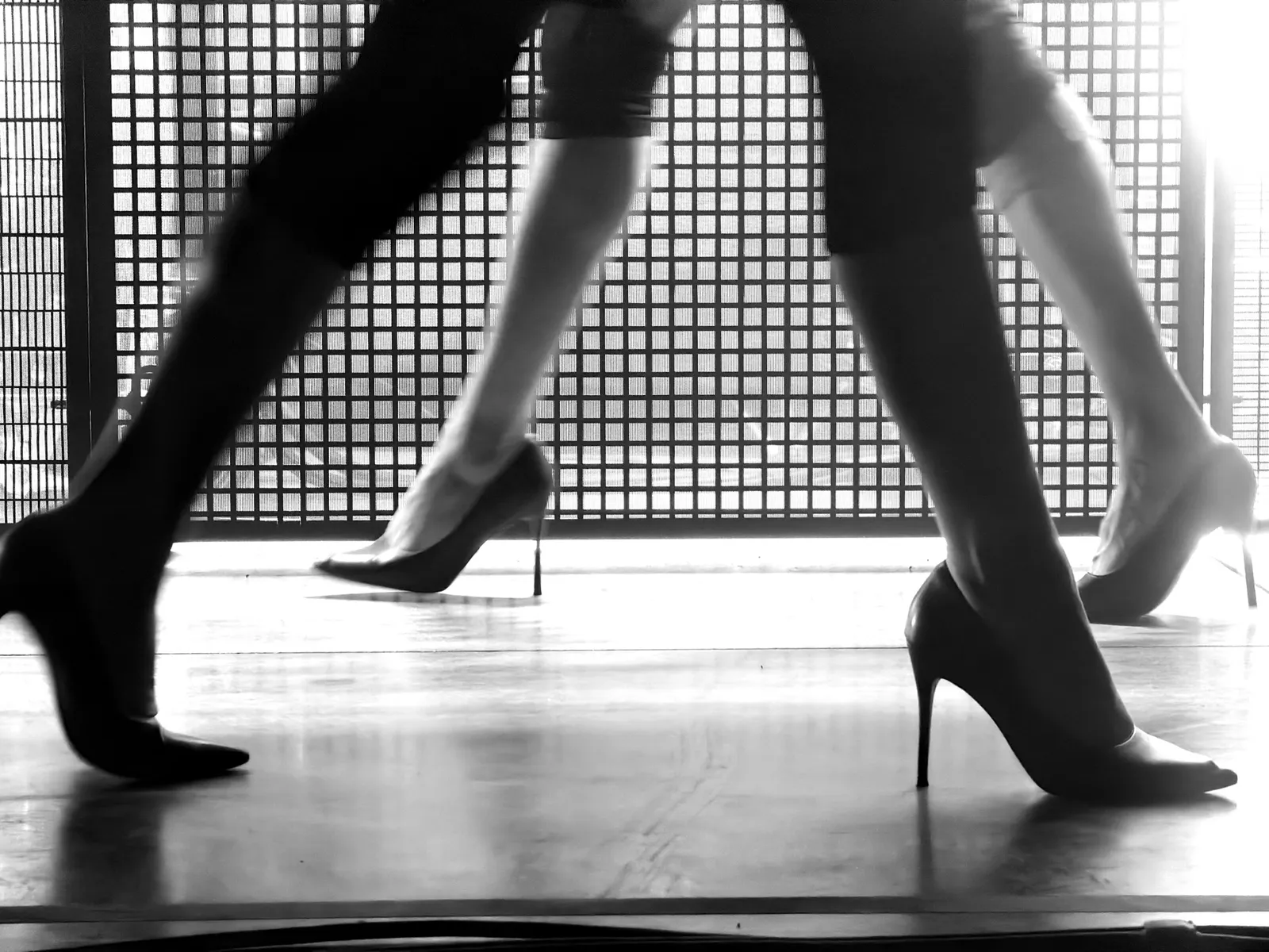 Black-and-white close-up of dancers walking in high heels across a brightly lit stage.