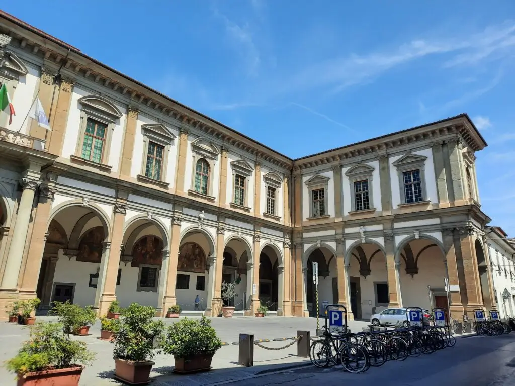 Historic Renaissance-style building with a columned arcade, symmetrical windows, and a sunlit courtyard lined with bicycles under a clear blue sky.