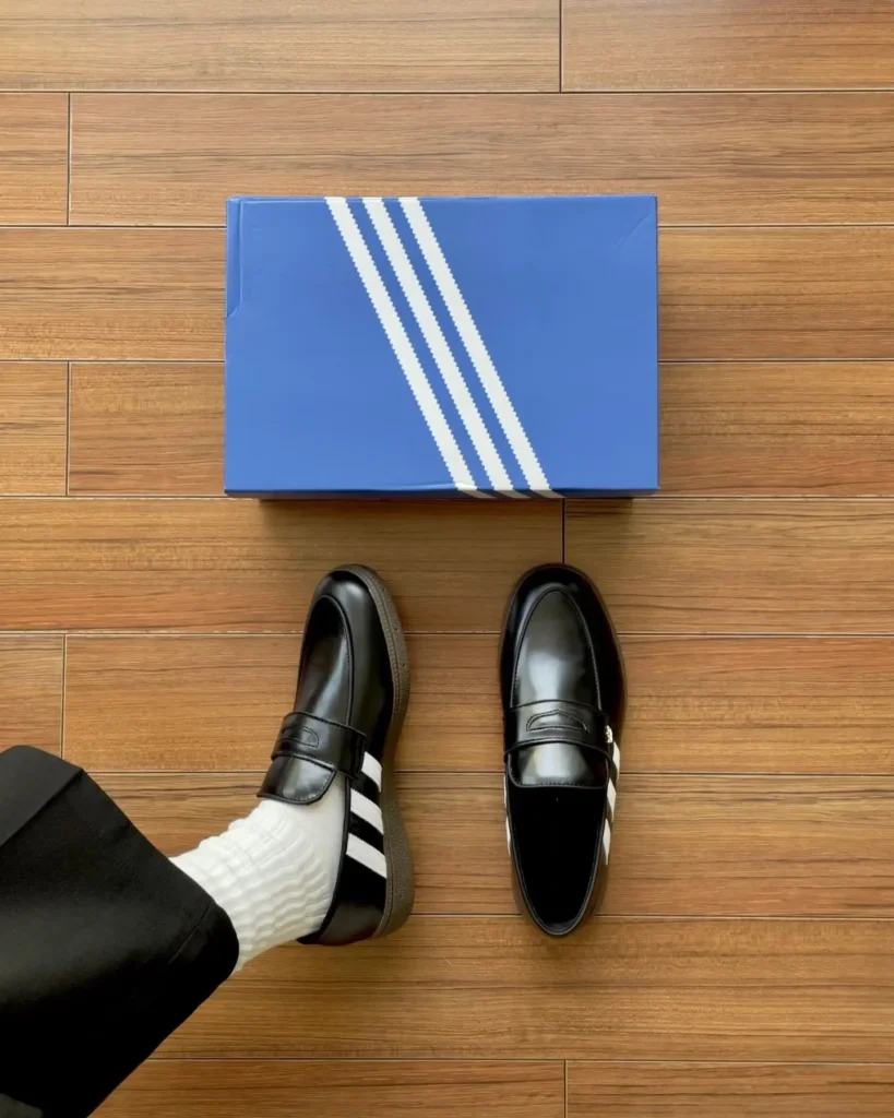 Overhead shot of a black leather adidas loafer hybrid with white three stripes worn on one foot, pair with the original blue and white Samba shoebox, on a wood floor