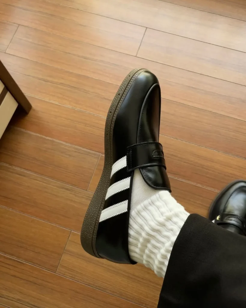 Close-up of a black leather loafer with white adidas three stripe and a gold trefoil logo on the strap, gum rubber sole view worn with a white sock