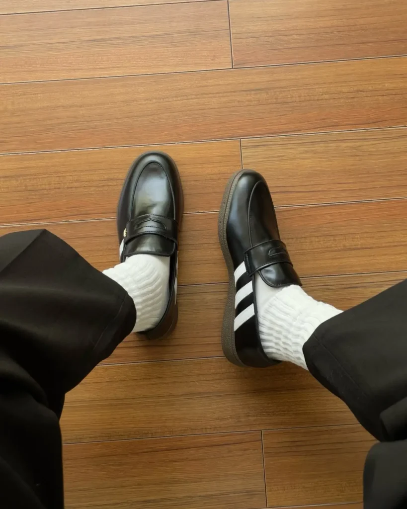 Overhead view of two feet wear black leather adidas loafer shoe with white stripe, white socks, and black trouser, on a wood floor