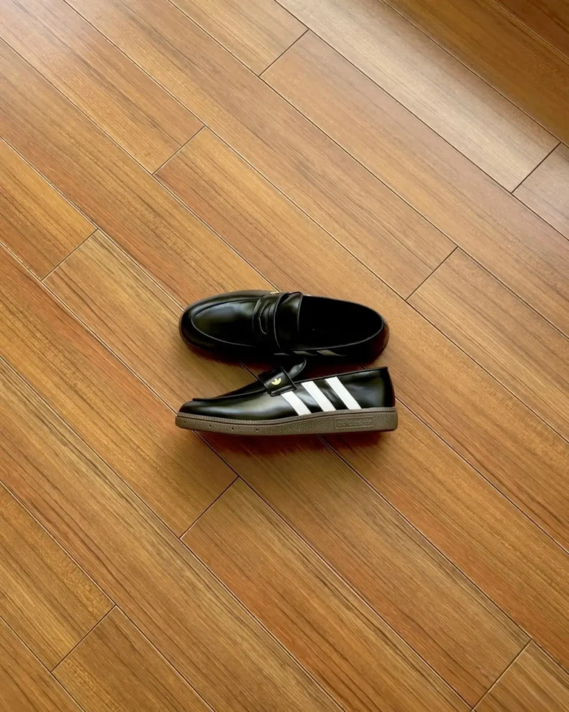 Pair of black leather Adidas loafer with white three stripes and gum soles, one shoe resting on top of the other on a wood floor