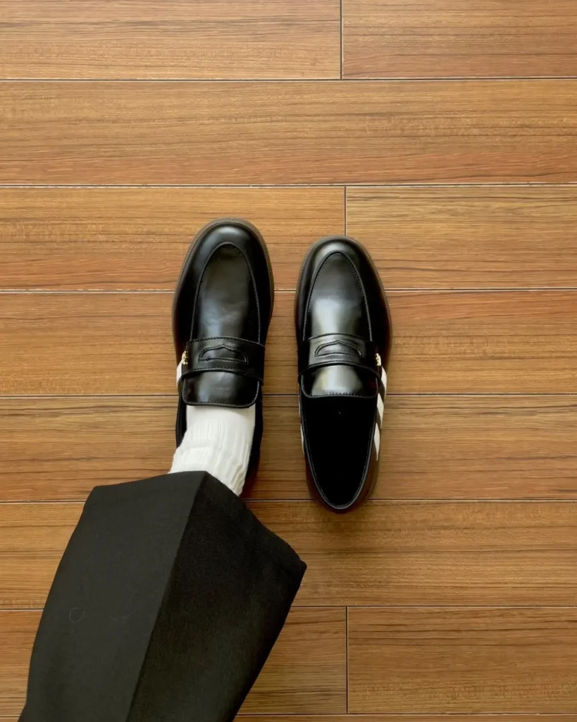 Front-facing overhead shot of black leather adidas loafer hybrid on a person feet, white ribbed sock, black trouser, wood floor background