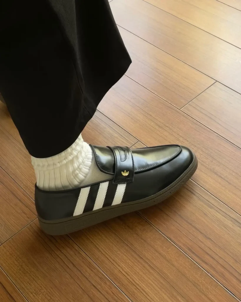 Overhead view of two feet wear black leather adidas loafer sneaker with white stripes, white socks, and black trouser, on a wood floor