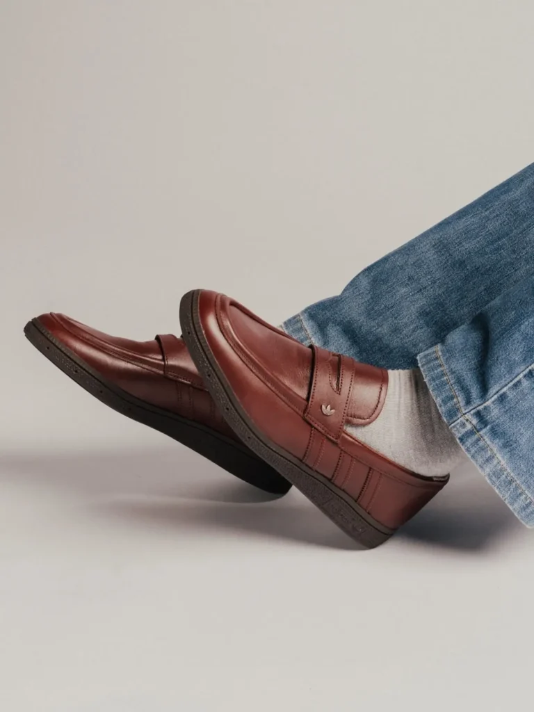 Studio product shot of a burgundy leather adidas loafer with tonal three stripe and trefoil logo, worn with grey sock and cuff jean against a plain grey backdrop