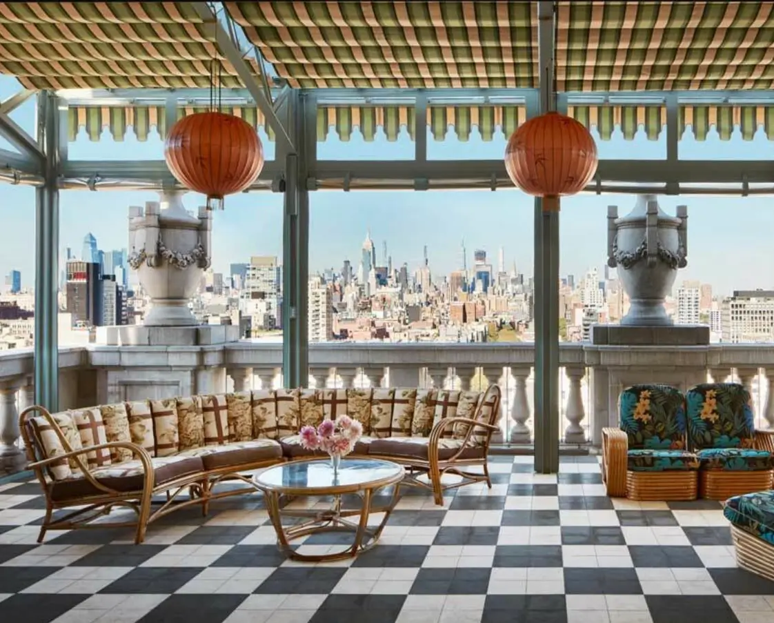 Rooftop terrace lounge overlooking the Manhattan skyline, featuring striped retractable awnings, red paper lanterns, rattan seating with patterned cushions, a black-and-white checkered tile floor, and classical stone balustrades framing the city view.