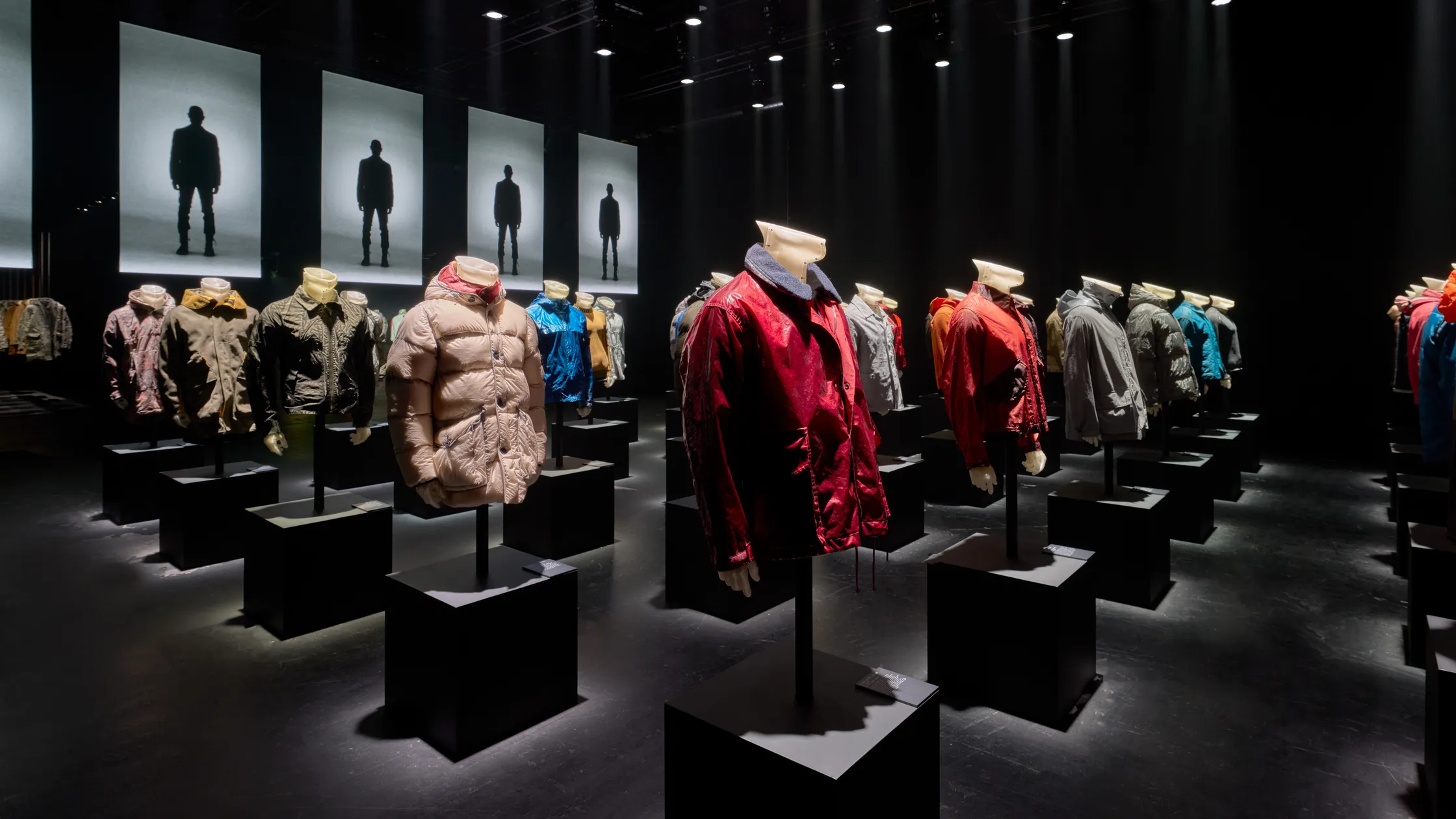 Stone Island exhibition installation featuring rows of colorful archival jackets displayed on headless mannequins atop black plinths in a dramatically lit gallery.