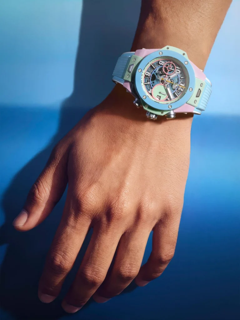 Hublot Big Bang watch sits on a wrist against a vivid blue background, feature a pastel blue bezel, mint and pink case, skeletonize dial, and blue strap