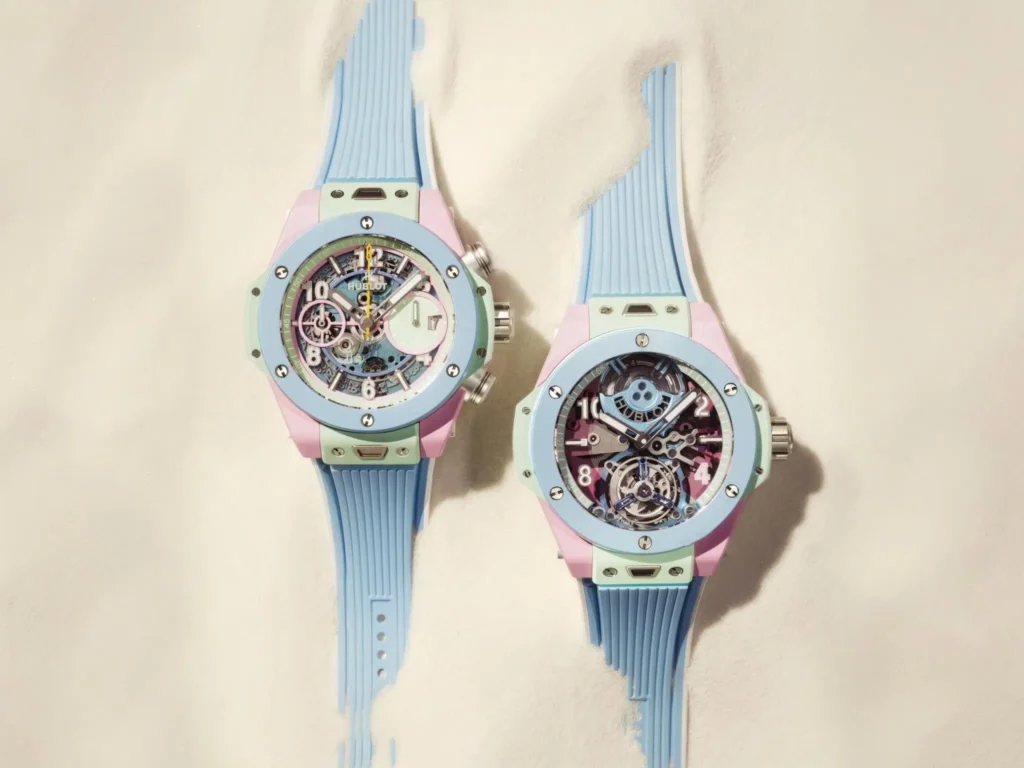 Two Hublot Big Bang watch lie side by side on a texture cream surface, each feature pastel blue, mint green, and pink case with blue strap and expo mech dial