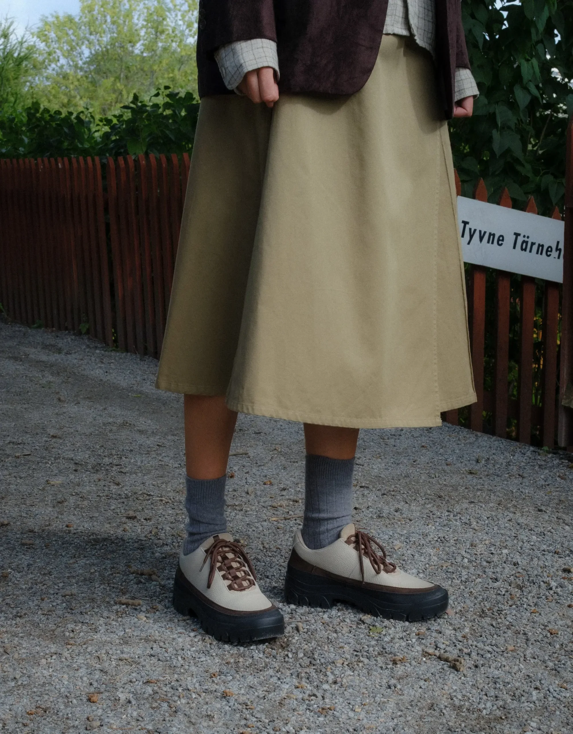 Person wearing off-white leather hiking shoes with brown suede accents, gray socks, and a beige skirt while standing on a gravel path.
