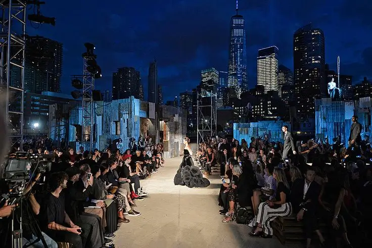 A nighttime fashion runway unfolds beneath the illuminated Manhattan skyline, with guests surrounding an industrial-inspired catwalk as models present dramatic evening looks.