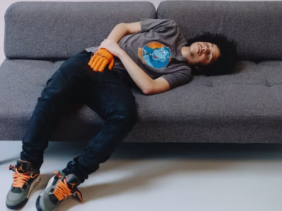 xaviersobase lounge on a gray sofa in a graphic T-shirt, black jeans, orange gloves, and gray-and-orange Air Jordan shoe