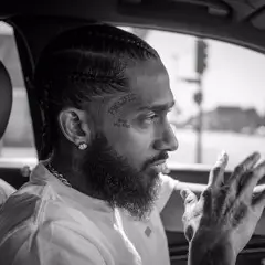 Black and white side profile of Nipsey Hussle with braids, beard, and facial tattoo in a car