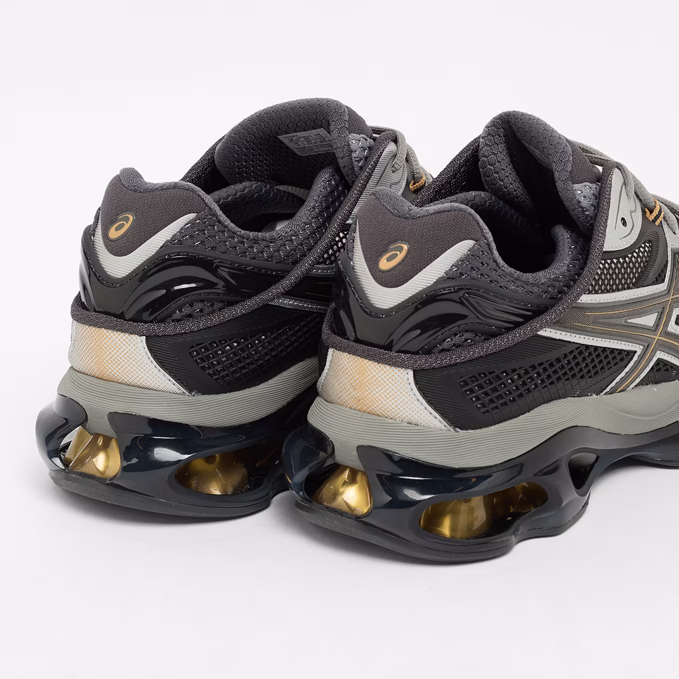 Rear three-quarter view of the ASICS GEL-KINETIC FLUENT in grey and black, showcasing glossy heel counters, layered mesh panels, and oversized visible GEL cushioning with metallic gold accents.
