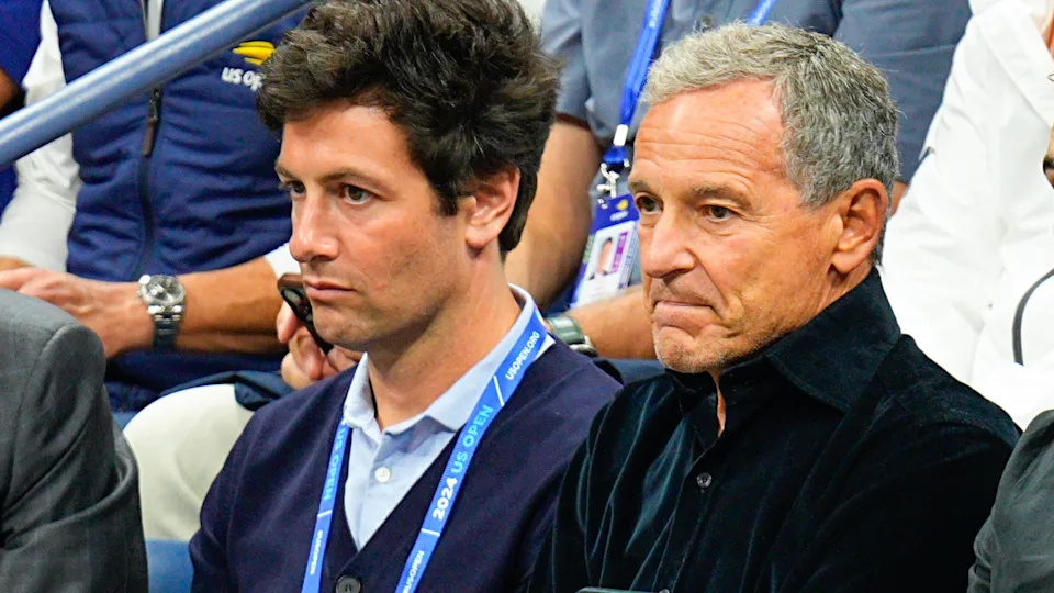 Two Men Sitting Courtside Together At The 2023 US Open, One Wearing A Navy Blazer And Tournament Lanyard And The Other Wearing A Black Jacket.