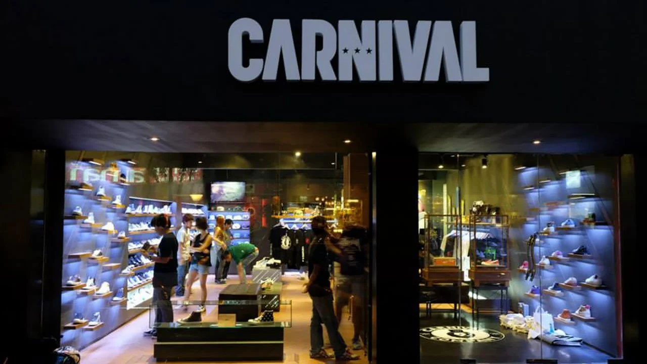 Storefront of the Carnival sneaker store at night, with an illuminated CARNIVAL sign above and shoppers browsing shelves of sneakers displayed under lit glass cases.