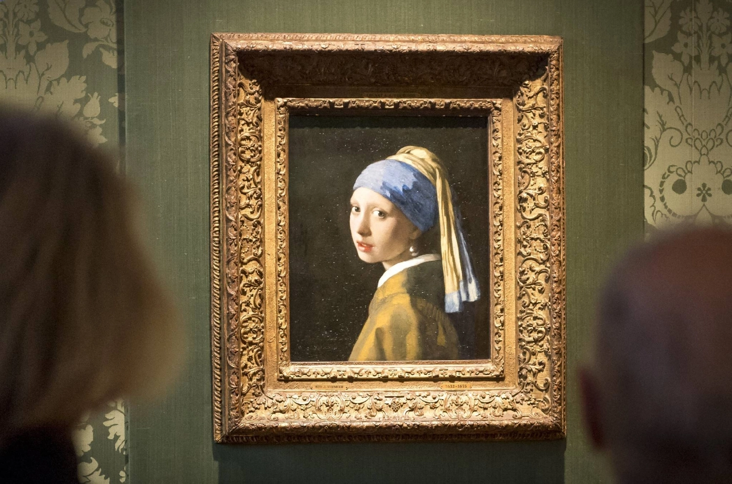 Johannes Vermeer's 'Girl with a Pearl Earring' displayed in an ornate gilded frame on a dark green museum wall, with blurred visitors in the foreground viewing the iconic Dutch masterpiece.