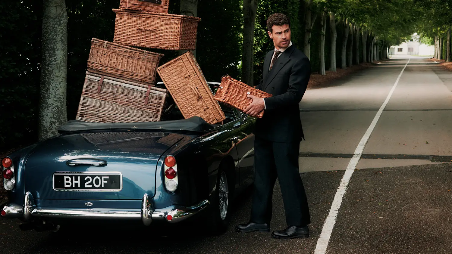 Man in a black suit and polished leather shoes loading wicker picnic baskets into a blue vintage convertible on a tree-lined road.