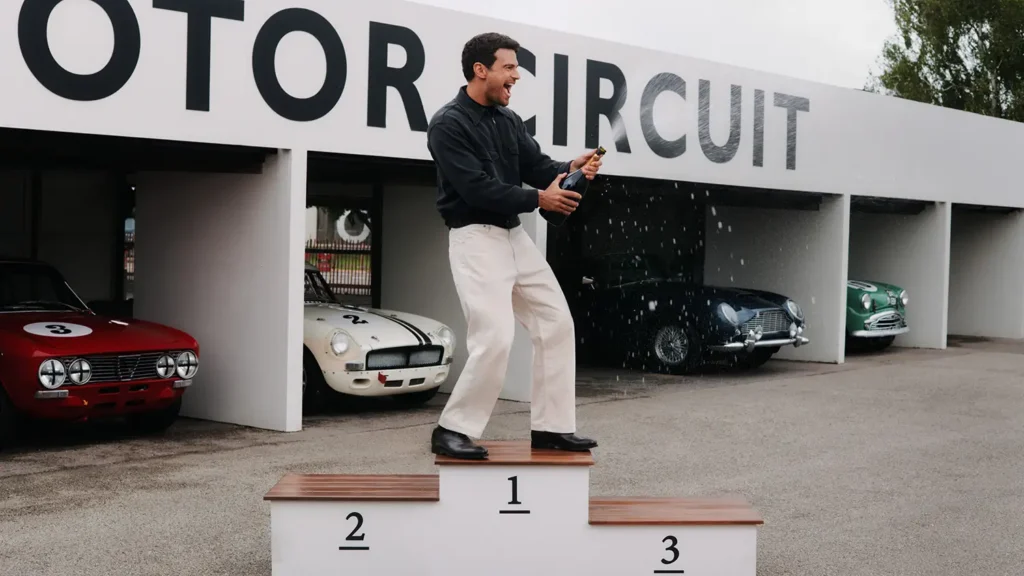 Man celebrate with a bottle on a winner podium while wear cream trouser and black leather shoe at motor circuit
