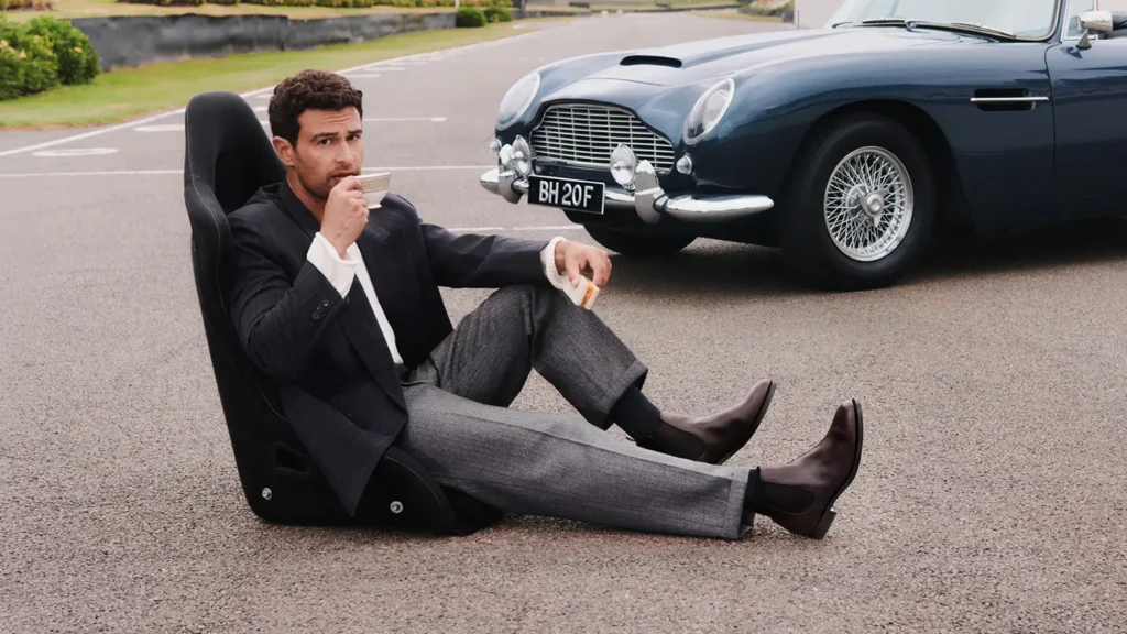 Seated man in tailor grey trouser and dark Chelsea boots drinking tea beside a blue vintage sport car
