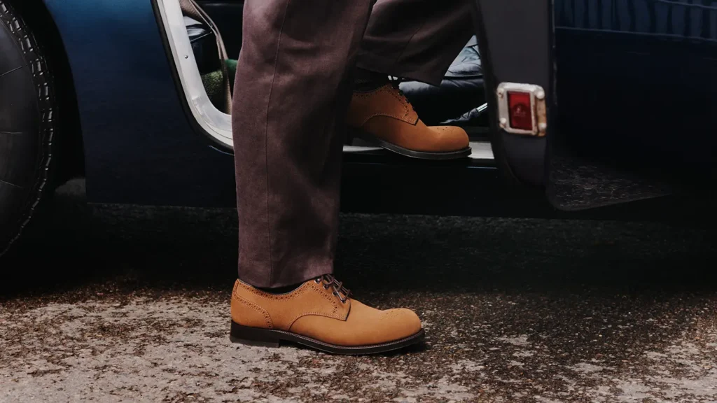 Tan suede Derby shoes paired with brown trousers beside the open door of a classic car
