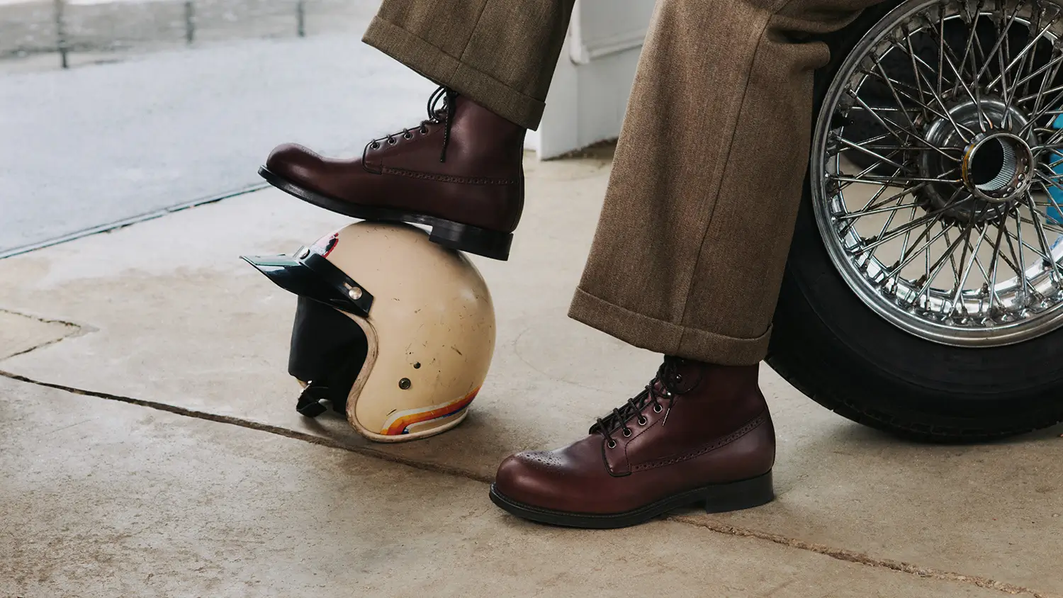 Dark brown leather lace-up boots worn with wide brown trousers beside a vintage racing helmet and wire-spoke car wheel.