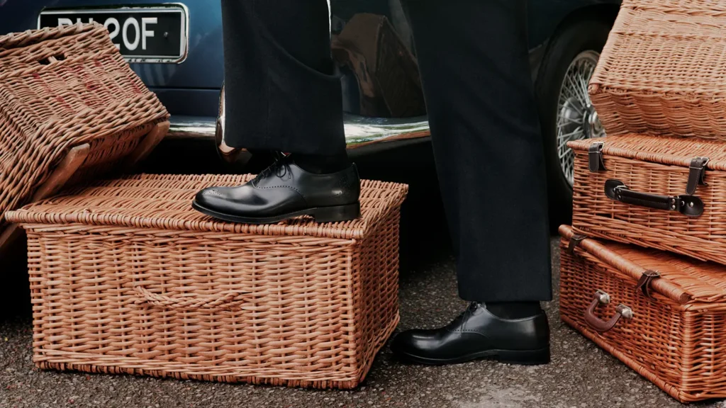 Polish black leather Oxford shoe worn with black trousers among stacked wicker picnic basket beside a classic car