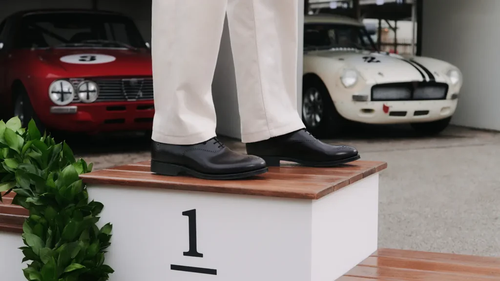 Black leather brogue shoes with cream trousers on a winner’s podium, with vintage race cars in the background