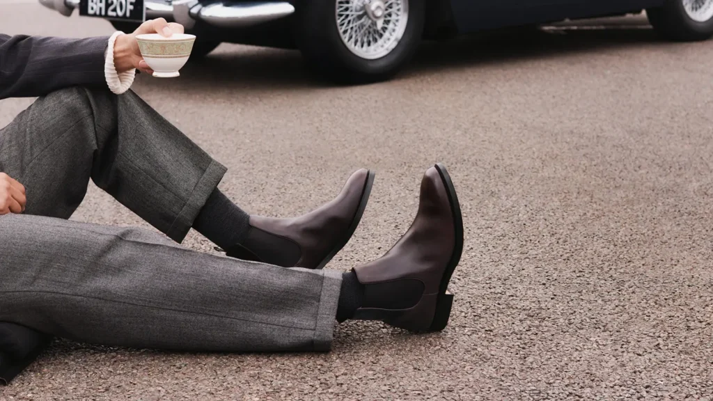 Dark brown Chelsea boots paired with grey tailored trousers as a seated man holds a teacup beside a classic car