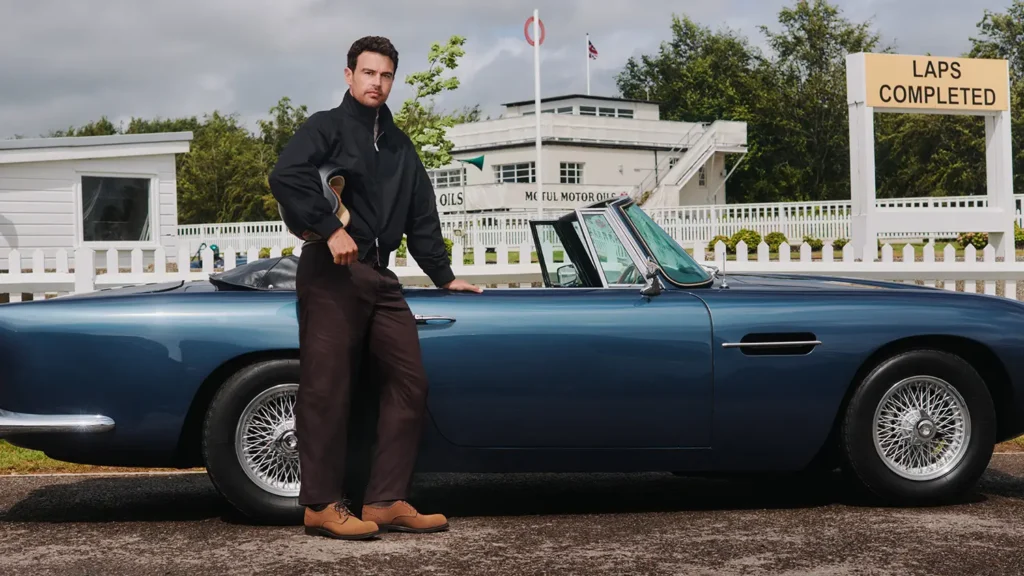 Man in brown trousers and tan Derby shoes leaning against a blue vintage convertible at a motor circuit