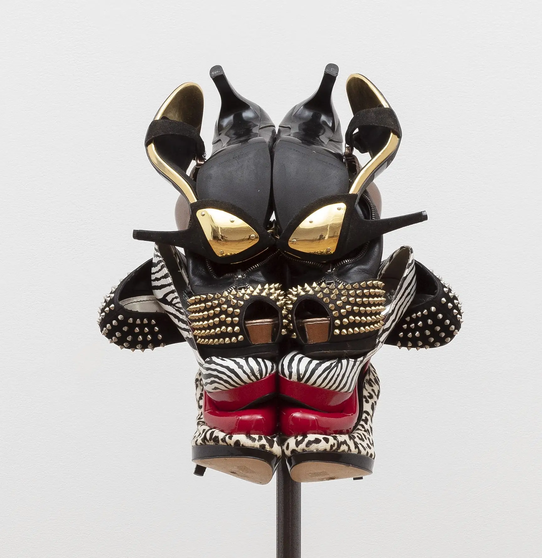 Contemporary sculptural artwork assembled from high-heeled shoes featuring black suede stilettos, zebra-print pumps, leopard-print heels, red soles, gold metallic accents, and studded leather arranged into a striking mask-like composition against a white background.