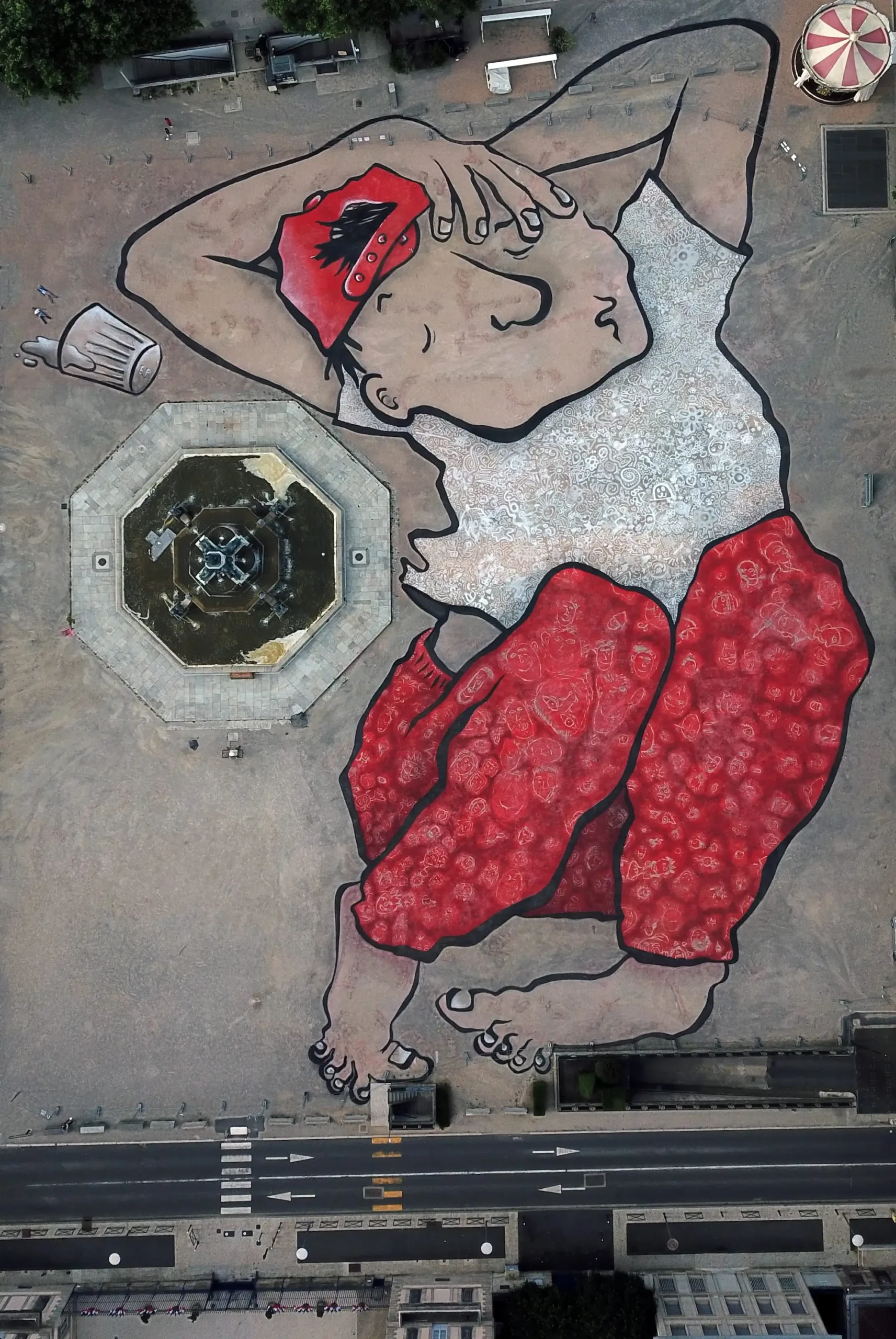 Aerial view of a monumental ground mural depicting a seated figure in red pants and cap beside an octagonal fountain.