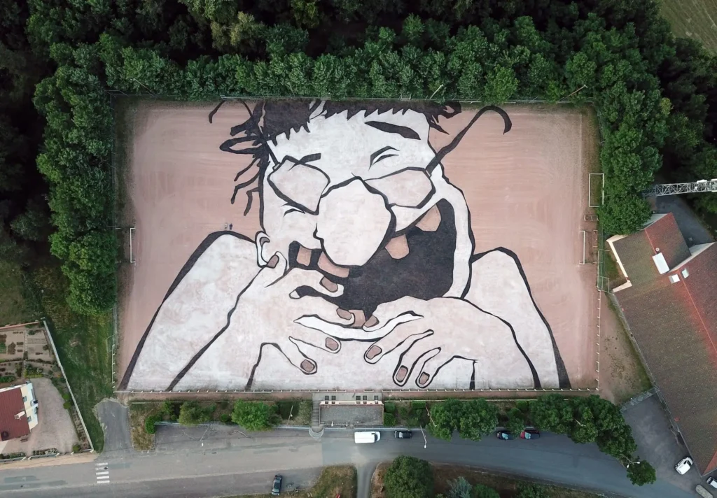 Aerial view of a massive black-and-white portrait painted across a sports field, depicting a laughing person with glasses and hands beneath their chin, surrounded by dense tree